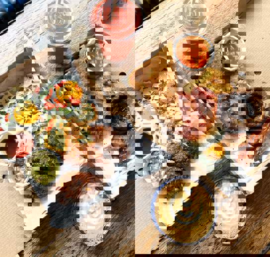 Two takeaway boxes filled with dishes. Our Sweetcorn Hash on the left and class Boss Breakfast on the right. A coffee and a smoothie in takeaway containers also sit on the table.