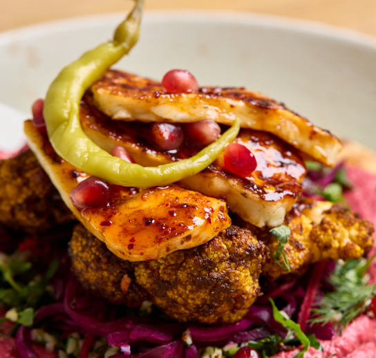 A close up shot of a flatbread with colourful houmous, slaw, cauliflower and halloumi on top with a thin, green chilli pepper
