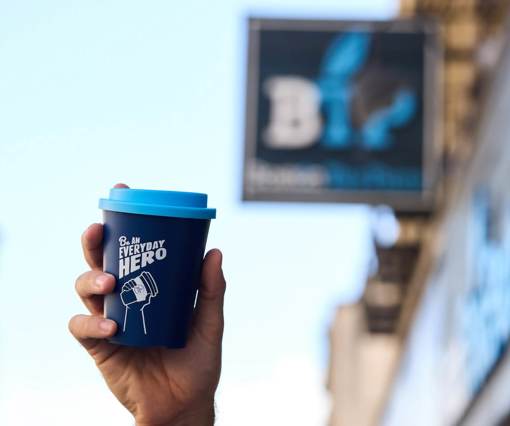 A hand holding a blue reusable coffee cup with the words 'Be an everyday hero' in white written across it