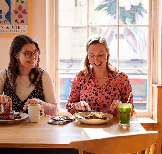 Two women, sitting next to one another, smiling and enjoying brunch dishes and drinks together